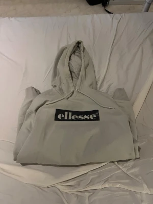 Beige Ellesse hoodie med logga - Snygg beige hoodie från Ellesse med svartvit logga på bröstet. Hoodien har huva med dragsko och en stor magficka framtill. Finns byxor om man vill köpa till.