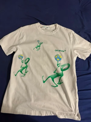 Vit Off-White t-shirt med grönt tryck - Vit t-shirt från Off-White med gröna tecknade figurer som jonglerar jordglober både fram och bak. Stort rött Off-White-kors på ryggen. Klassisk passform och rund hals. Perfekt för dig som gillar streetwear och unika prints.