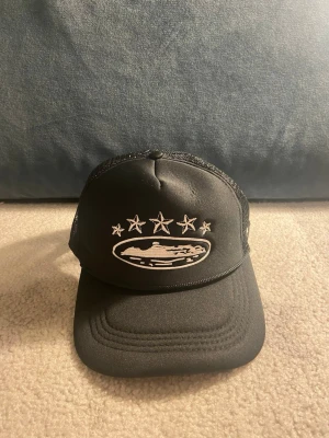 Svart truckerkeps med stjärnor - Snygg svart truckerkeps med mesh på sidorna och broderade vita stjärnor framtill och på sidan. Framtill finns även ett broderat motiv av ett ufo i vitt. Justerbar baktill för perfekt passform. Perfekt för dig som gillar streetwear och unika detaljer.