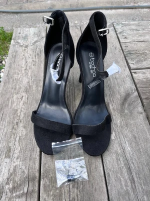 Svarta klackpumps från Boohoo - Helt oanvända, med pappret och plasten kvar.  Stilrena svarta pumps från Boohoo med smal rem över tårna och justerbar ankelrem med silverfärgat spänne. Skorna har hög smal klack och ovandel i mockaimitation. Perfekta för att lyfta din outfit med en elegant touch. Storlek 39