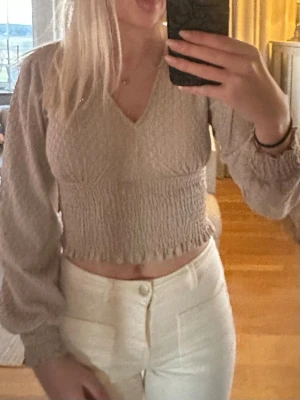 Beige croppad blus med v-ringning - Snygg beige croppad blus med v-ringning och långa puffärmar. Toppen har smockad midja och ärmar med resår vid handlederna. Perfekt för dig som gillar en feminin och trendig stil.