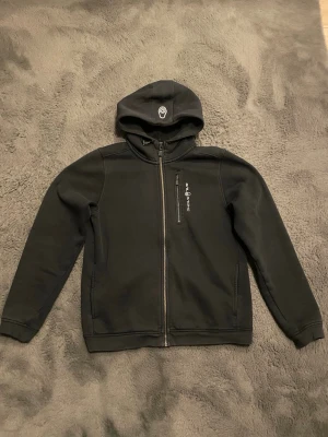 Svart hoodie från Sail Racing - Snygg svart hoodie från Sail Racing med dragkedja framtill och broderad logga på huvan. Tillverkad i mjukt material med fleecefoder på insidan för extra komfort. Storleken är S / 176
