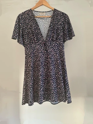 Blommig kortklänning från Pull&Bear i S - Söt kortklänning från Pull&Bear med mörkblå bas och små vita och rosa blommor. Klänningen har v-ringning, korta ärmar och ett lätt, flödande material som ger en avslappnad och feminin look. Perfekt för dig som gillar blommiga mönster och enkel stil.
