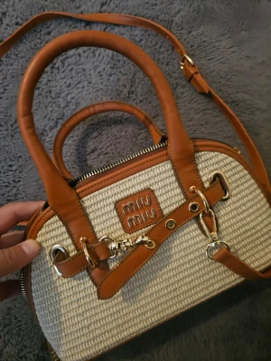 Miu Miu beige och brun handväska - Snygg handväska från Miu Miu i beige flätad material med bruna läderdetaljer och guldfärgade metalldelar. Väskan har både handtag och axelrem samt ett dekorativt spänne framtill. Perfekt för dig som gillar trendiga accessoarer.
