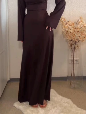 Brun långklänning med snörning - Elegant brun långklänning med långa ärmar och snörningsdetalj i midjan. Klänningen hat en figurnära passform upptill och faller snyggt längs kroppen. Perfekt för dig som gillar stilrena och tidlösa plagg.
