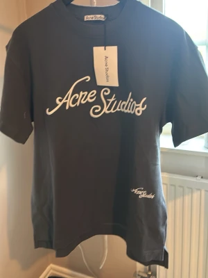 Mörkgrå  t-shirt från Acne Studios - Mörkgrå t-shirt från Acne Studios med vit logotyp framtill i snyggt tryck. Klassisk passform med rund hals och korta ärmar. Tillverkad i mjuk bomull, perfekt för en stilren och avslappnad look.