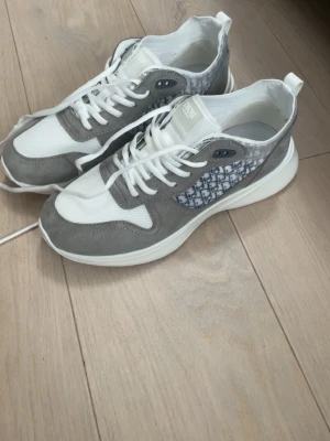 Gråvita sneakers med monogram - Snygga sneakers från Zara  i grått och vitt med klassiskt monogram på sidan. Skorna har en chunky vit sula, ovandel i mocka och mesh samt vita skosnören. Perfekta för dig som vill ha en stilren och exklusiv look.