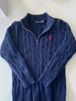 Mörkblå kabelstickad tröja Polo Ralph Lauren - Mörkblå kabelstickad tröja från Polo Ralph Lauren med halv dragkedja och hög krage. Tröjan har ett broderat rött logomärke på bröstet och ribbstickade muddar. Perfekt för lager-på-lager och en klassisk look.