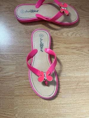 Rosa sandaler med fjärilsdetalj - Snygga rosa sandaler från Chatties med en söt fjärilsdetalj i metall framtill. Sandalerna har en klassisk flip-flop design med breda remmar i plast och en beige innersula. 