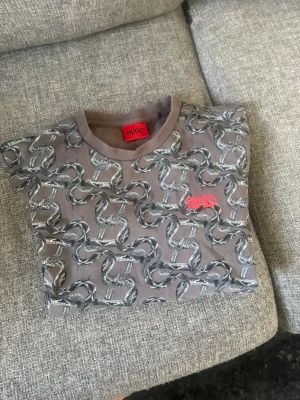 Grå mönstrad sweatshirt från Hugo - Säljer en grå sweatshirt från Hugo med coolt ormmönster över hela tröjan och röd Hugo-logga på bröstet. Tröjan har rund halsringning, långa ärmar och ribbade muddar. Perfekt för dig som vill sticka ut med en unik design.
