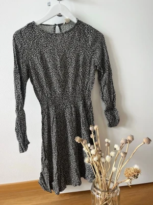 Svartvit blommig klänning med lång ärm - Svart klänning med vita småblommiga mönster och lång ärm. Modellen är kort och har smock i midjan för en snygg passform. Tillverkad i ett mjukt och lätt material som känns skönt mot huden. Perfekt för dig som gillar en feminin och trendig stil.