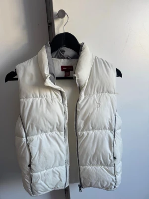 Vit dunväst från H&M - Snygg och puffig vit dunväst från H&M, hög krage och dragkedja framtill. Västen har två sidofickor och diskret logga på bröstet. Perfekt för lager-på-lager och håller dig varm under kyliga dagar.