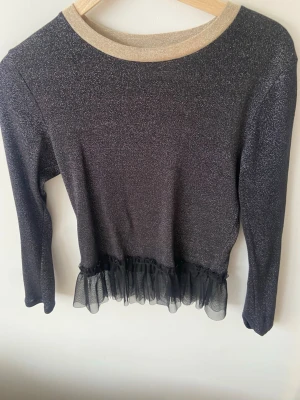 Glittrig svart topp med volang från MADS NORGAARD - Snygg svart topp med glittrigt tyg och beige krage. Toppen har lång ärm och en transparent volang i mesh längst ner för extra detalj. Perfekt för fest!