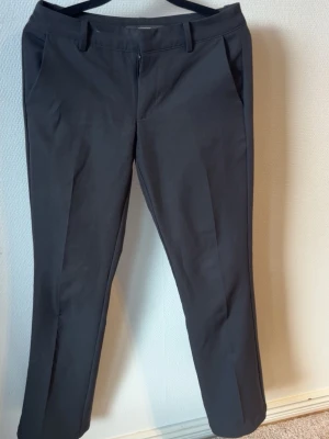 Svarta kostymbyxor med raka ben - Snygga svarta kostymbyxor med raka ben och pressveck. Byxorna har två sidofickor fram och två passpoalfickor bak. Klassisk midja med hällor för skärp och diskret dragkedja. Perfekta till en stilren look.