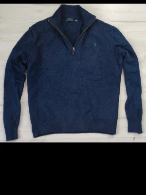 Blå halvzip tröja från Polo Ralph Lauren - Mörkblå stickad tröja från Polo Ralph Lauren med halv dragkedja och hög krage. Tröjan har diskret logga broderad på bröstet och ribbade muddar vid ärmslut och nederkant. Perfekt för lager på lager och en clean look.