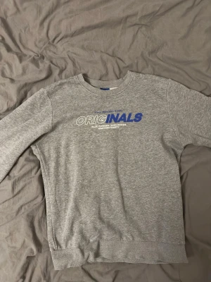 Grå sweatshirt från Jack & Jones - Grå sweatshirt från Jack & Jones Originals med tryck i vitt och blått framtill. Tröjan har rund hals, långa ärmar och ribbade muddar vid ärmslut och nederkant. Tillverkad i mjukt bomullsmaterial för en bekväm känsla.