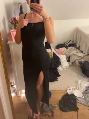 Svart långklänning med slits - Säljer en svart långklänning med smala axelband och en snygg slits framtill. Klänningen har en figurnära passform och enkel, stilren design som ger en elegant look. Perfekt för dig som vill ha något klassiskt men ändå trendigt.