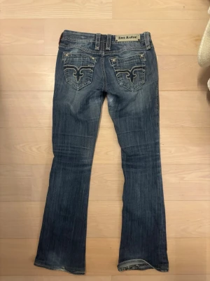 Rock Revival bootcut jeans blå - Säljer ett par blå bootcut jeans från Rock Revival med snygga slitningar och broderade detaljer på bakfickorna. Jeansen har låg midja och klassisk femficksmodell. Perfekta för dig som gillar en avslappnad men trendig stil.