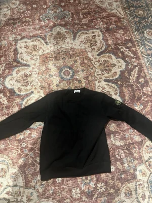 Stone Island tröja - Svart sweatshirt från Stone Island med klassisk rund hals och ribbade muddar. Tröjan har den ikoniska Stone Island-loggan på vänster ärm. Perfekt för dig som gillar stilrena och tidlösa plagg.
