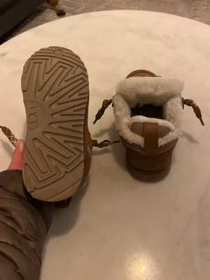 Bruna UGG boots med fårskinn - Säljer ett par bruna kopia UGG boots med snörning och mjukt vitt fårskinnsfoder i färgen chestnut. Skorna har en  gummisula med UGG-logga undertill och är perfekta för kalla dagar. Klassisk design med låg skaft och dragflik bak. Köpte dom och har använt dem tre gånger men de var för små.