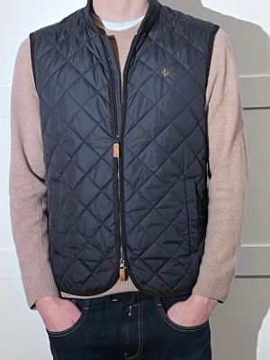 Marinblå quiltad väst från Morris - Snygg marinblå quiltad väst från Morris med diskret logga på bröstet. Västen har dragkedja framtill, ståkrage och två sidofickor. Klassisk passform och rutmönstrad design gör den enkel att matcha till olika outfits.