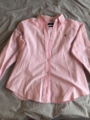 Rosa skjorta från Ralph Lauren - Klassisk rosa skjorta från Ralph Lauren med broderad logga på bröstet. Skjortan har lång ärm, knappar framtill och är tillverkad i 100% bomull. Perfekt för en clean och stilren look.