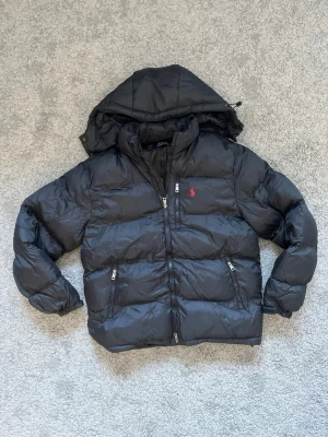 Ralph lauren puffer jacket - Säljer en svart pufferjacka från Polo Ralph Lauren med avtagbar huva och två dragkedjefickor framtill. Jackan har det klassiska röda Polo-logot på bröstet och breda kanaler för extra värme. Perfekt för kalla vinterdagar.