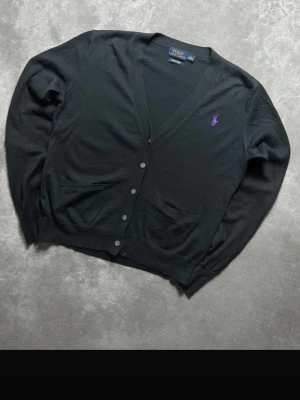 Svart cardigan från Polo Ralph Lauren - Klassisk svart cardigan med V-ringning från Polo Ralph Lauren. Tröjan är gjord av 100% pima bomull. Tröjan har knappstängning framtill, två fickor och en lila broderad logga på bröstet. Perfekt att slänga över en t-shirt för en clean look. Skick 10/10. Sjukt limiterad second hand i det här skicket. Otroligt fin utan hål eller andra defekter vid frågor eller funderingar är det bara att säga till Mvh Olof.