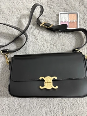 Svart axelväska  - Snygg svart axelväska. i slätt läder med justerbar axelrem och ikoniskt guldigt metallspänne framtill. Väskan har en rektangulär form och minimalistisk design som ger en lyxig känsla. Perfekt för dig som gillar stilrena accessoarer.