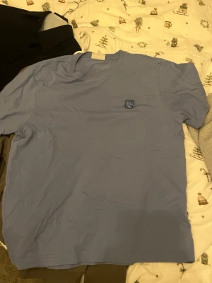 Blå t-shirt från Kitsune - Snygg blå t-shirt med ett litet vargmärke broderat på bröstet. T-shirten har en klassisk passform och rund halsringning, perfekt för en avslappnad stil. Tillverkad i mjuk bomull som känns skön mot huden.