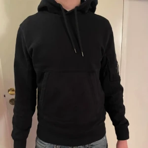 Svart hoodie från C.P. Company - Svart hoodie från C.P. Company med klassisk huva och snörning, stor magficka och ikonisk linsdetalj på ärmen. Tröjan är i mjuk bomull och har en clean, stilren look som passar perfekt till jeans eller joggers.