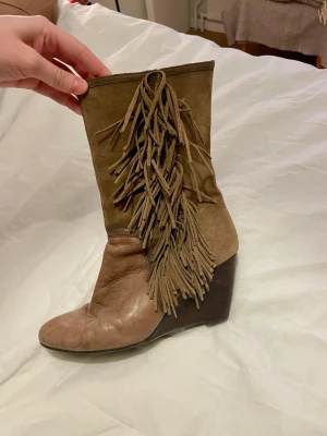 Bruna cowboy boots med fransar - Säljer ett par bruna cowboy boots med kilklack och långa fransar på skaftet. Bootsens ovandel är i mocka och fotdelen i slätt läder, vilket ger en snygg kontrast. De har en cool westernstil och är perfekta för dig som vill sticka ut.