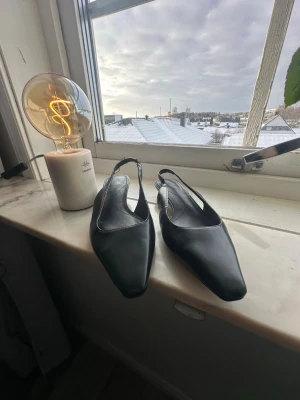 Svarta ballerinaskor med fyrkantig tå - Stilrena svarta ballerinaskor från H&M med öppen häl och fyrkantig tå. Skorna har en tunn rem runt hälen och är tillverkade i ett slätt material som ger en modern look. Perfekta för dig som gillar minimalistisk stil. Väl andvända!!