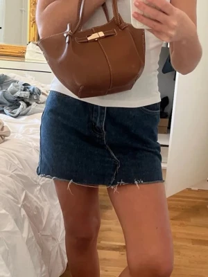 Mörkblå jeanskjol med rå kant - Trendig mörkblå jeanskjol med rå, fransig kant nertill och klassisk femficksdesign. Kjolen har en rak passform och är perfekt för en avslappnad look. Passar till både sneakers och boots.