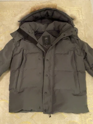 Grå dunjacka från Canada Goose - Snygg grå dunjacka från Canada Goose med avtagbar huva och pälskant. Jackan har dragkedja och knappar framtill, två stora fickor och en hög krage. Perfekt för kalla vinterdagar och riktigt varm tack vare dunfyllningen.