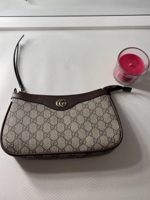  beige monogram handväska - Snygg beige handväska med klassiskt GG-monogram i canvas och mörkbruna läderdetaljer. Väskan har en elegant guldtonad logga framtill och smal axelrem i läder. Perfekt för dig som gillar lyxiga accessoarer och tidlös design.