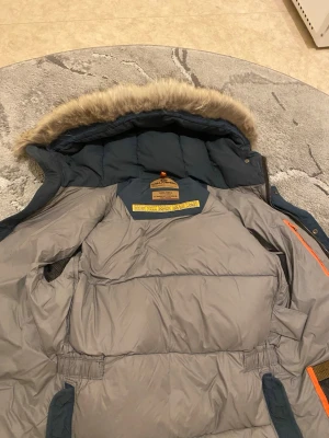 Mörkblå dunjacka från Parajumpers - Snygg mörkblå dunjacka från Parajumpers med avtagbar fuskpäls på huvan och orange detaljer. Jackan har hög fyllnadsgrad med 90/10 dun/fjäder och är perfekt för kalla vinterdagar. Insidan är grå och jackan har flera praktiska fickor.