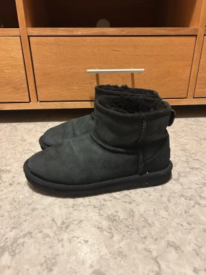 Svarta UGG boots i mocka - Ett par svarta UGG boots i mjuk mocka med kort skaft och varm, fluffig insida i fårskinn. Klassisk UGG-sula med logga undertill och enkel, stilren design. Perfekta för kalla dagar när du vill ha både stil och komfort.