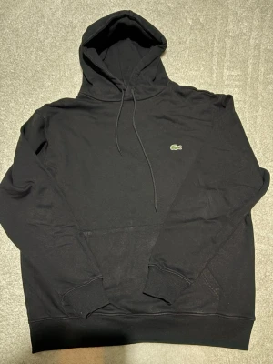 Svart hoodie från Lacoste - Säljer en svart hoodie från Lacoste med klassisk krokodillogga på bröstet. Hoodien har huva med dragsko, känguruficka och ribbade muddar vid ärmar och nederkant. Tillverkad i mjukt bomullsmaterial, perfekt för en chill och stilren look.