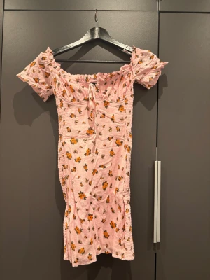 Rosa blommig kortklänning med snörning - Söt rosa kortklänning med små blommor i orange och grönt. Klänningen har offshoulder-ärmar men går att ha som vanligt, figurnära passform och en liten snörning framtill. Lite skrynklig pga legat länge. 