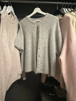 Grå ribbstickad kofta med kort ärm - Mysig grå ribbstickad kofta med korta ärmar och rund hals. Koftan har knappar framtill i en ljus nyans och är tillverkad i ett mjukt, fluffigt material som ger en avslappnad look. Perfekt att slänga över en topp för extra lager.