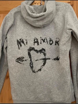 Grå hoodie Mi Amor - Grå hoodie med huva och svart tryck på ryggen där det står 'Mi Amor' och ett hjärta genomborrat av en pil. Hoodien har en avslappnad passform och är tillverkad i mjukt sweatshirtmaterial. Perfekt för en chill och trendig look.