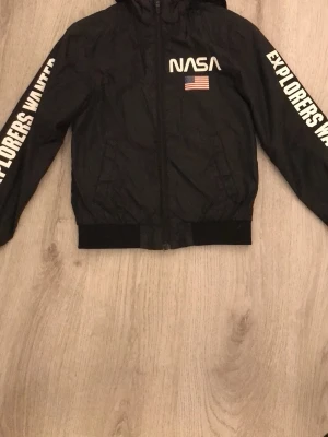 Svart NASA vindjacka med tryck - Svart vindjacka med dragkedja, huva och resår i midja och ärmslut. Stort NASA-tryck och amerikansk flagga på bröstet samt texten 'EXPLORERS WANTED' längs båda ärmarna. Lätt material, perfekt för blåsiga dagar.