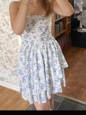 Vit blommig axelbandslös klänning - Söt axelbandslös klänning i vitt med blått blommönster. Klänningen har en volangig kjol och sitter åt upptill. Perfekt för varma dagar och har en romantisk vibe.