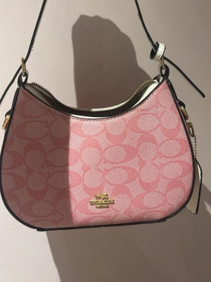 Rosa axelremsväska från Coach - Snygg rosa handväska från Coach med klassiskt monogrammönster i tyg. Väskan har guldfärgade detaljer, dragkedja upptill och justerbar axelrem i svart och vitt.  Har många rosa väskor Darför säljer jag denna , skick 10/10