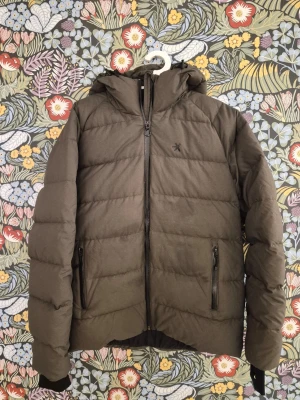 Olivgrön pufferjacka med huva från Everest M - Snygg olivgrön pufferjacka med huva och svarta dragkedjor. Jackan har två framfickor med dragkedja och quiltad design. Perfekt för kalla dagar och har ribbade muddar vid ärmsluten. Liten diskret logga på bröstet.