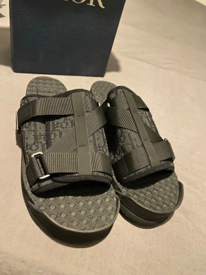 Svarta Dior sandaler med monogram - Säljer ett par svarta sandaler från Dior med tydligt monogrammönster och breda justerbara remmar i textil. Sulan har prickigt mönster för extra grepp och komfort. Perfekta för dig som vill ha en lyxig och trendig look.