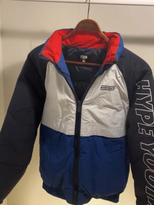 FSBN Hype Youth pufferjacka blå/vit - Säljer en snygg pufferjacka från FSBN med Hype Youth-tryck. Jackan har färgblock i blått, vitt och svart samt röd insida på kragen. Dragkedja framtill och elastiska muddar. Perfekt för kalla dagar och riktigt streetig stil.
