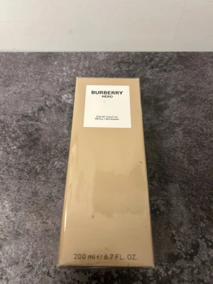 Burberry Hero Eau de Toilette 200 ml Äkta - Burberry Hero Eau de Toilette i en stilren beige kartong med minimalistisk design. Flaskan innehåller 200 ml och är en fräsch och maskulin doft. Förpackningen har tydlig märkning och klassisk Burberry-känsla. Perfekt för dig som gillar exklusiva parfymer.