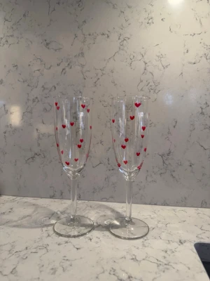 Champagneglas med röda hjärtan - Två eleganta champagneglas i genomskinligt glas dekorerade med små röda hjärtan runt hela kupan. Perfekta för speciella tillfällen eller som en söt detalj i köket.